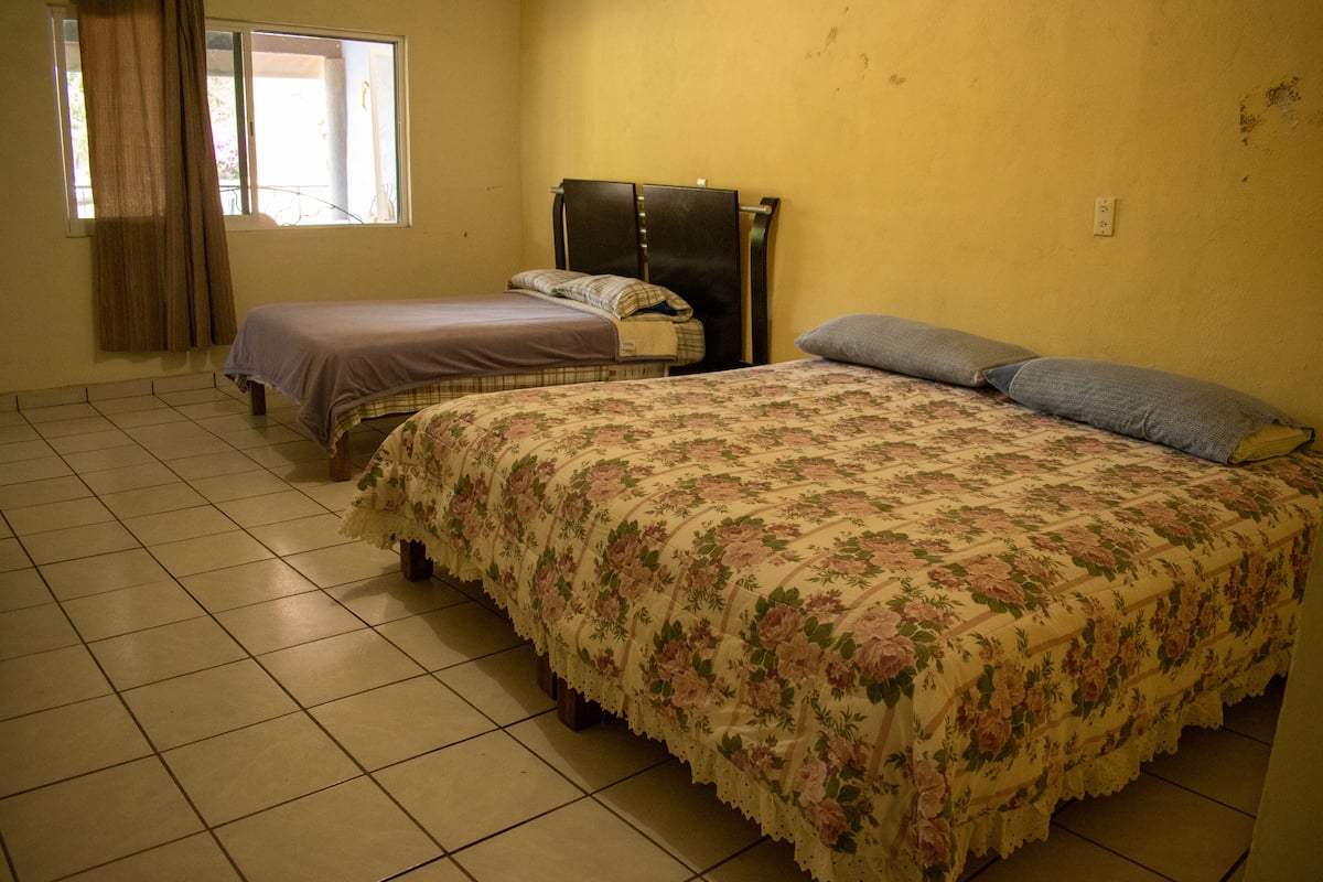 Habitación con dos camas, una con colcha estampada y la otra con cubierta lisa. Hay una ventana con cortinas, un enchufe de pared y piso de baldosas.