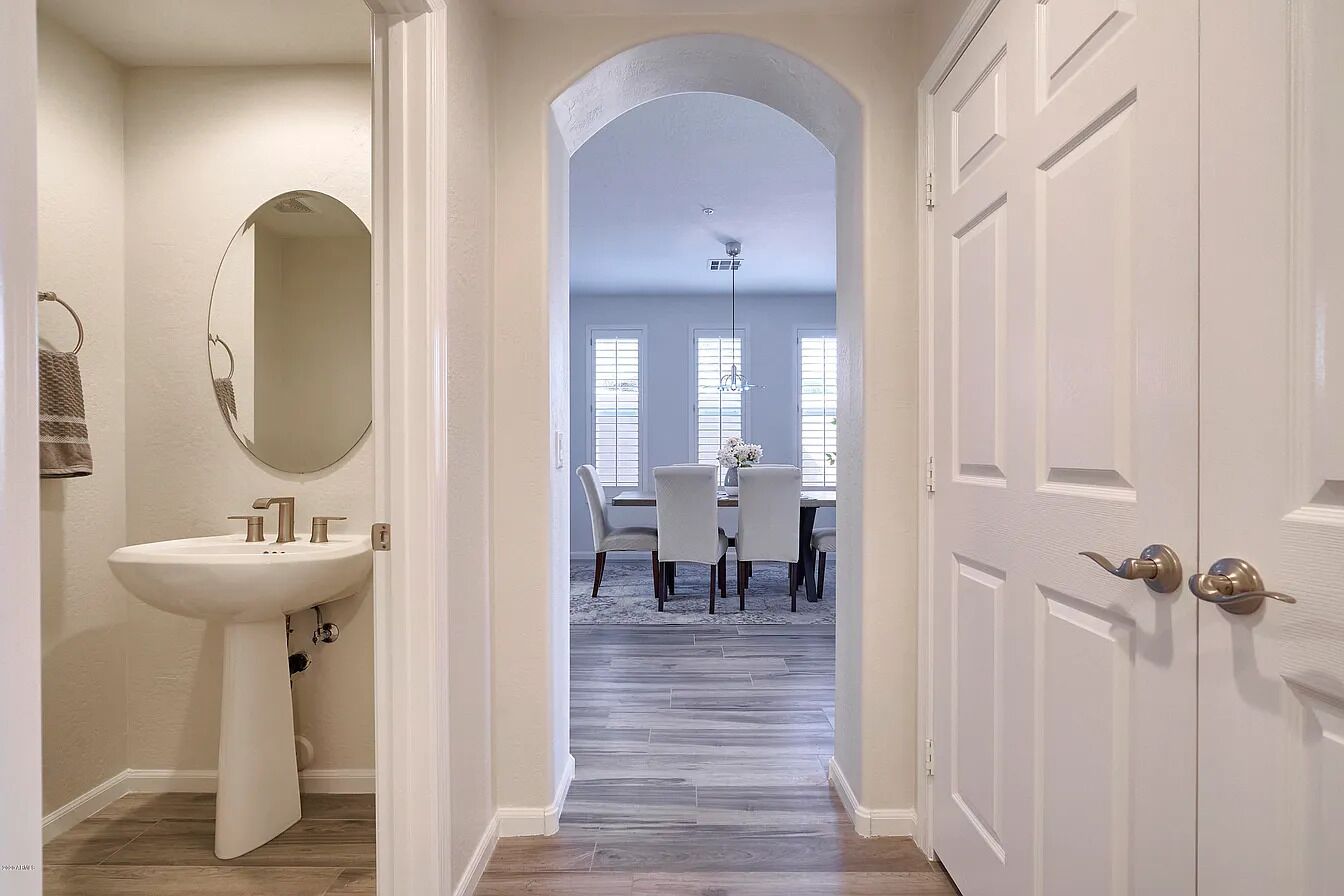 A hallway leading to a dining area with a white sink and mirror, a chandelier, and a dining table with chairs.