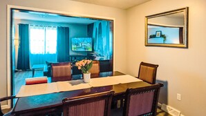 Un coin repas avec une table, des chaises et un vase de fleurs. On aperçoit un téléviseur au fond et un miroir est accroché au mur.