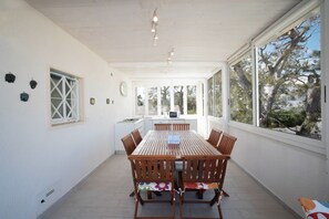A spacious dining area with a wooden table and chairs, large windows offering views of trees, and a kitchen in the background.