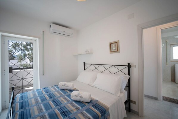 A bedroom with a bed, white pillows, a blue and beige striped bedspread, an air conditioning unit, a framed picture on the wall, and a door leading to another room.
