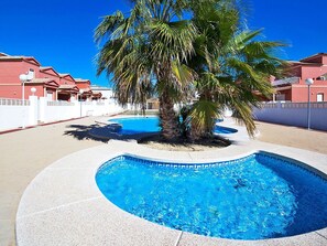 A swimming pool with a palm tree in the center, surrounded by a white fence and red buildings in the background.