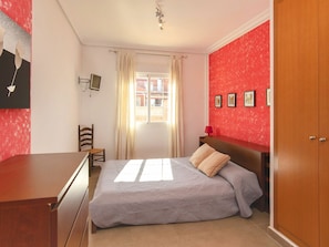 A bedroom with a bed, a desk, a chair, a window with curtains, a TV, and a red wall with framed pictures.