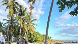 Ein Essbereich am Strand mit weißen Stühlen und Tischen, Palmen und freiem Blick auf das Meer.