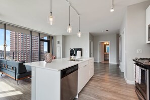 A modern kitchen with a dishwasher, a sink, and a large island. There is a dining area with a sofa and a large window offering a city view.