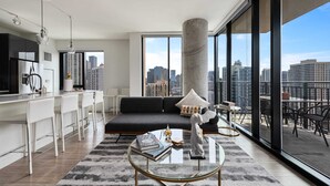 Ein modernes Wohnzimmer mit einer schwarzen Couch, einem Glastisch und Blick auf die Stadt durch große Fenster.