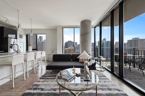 Un salón moderno con un sofá negro, una mesa de centro de vidrio y vistas a la ciudad a través de amplios ventanales.