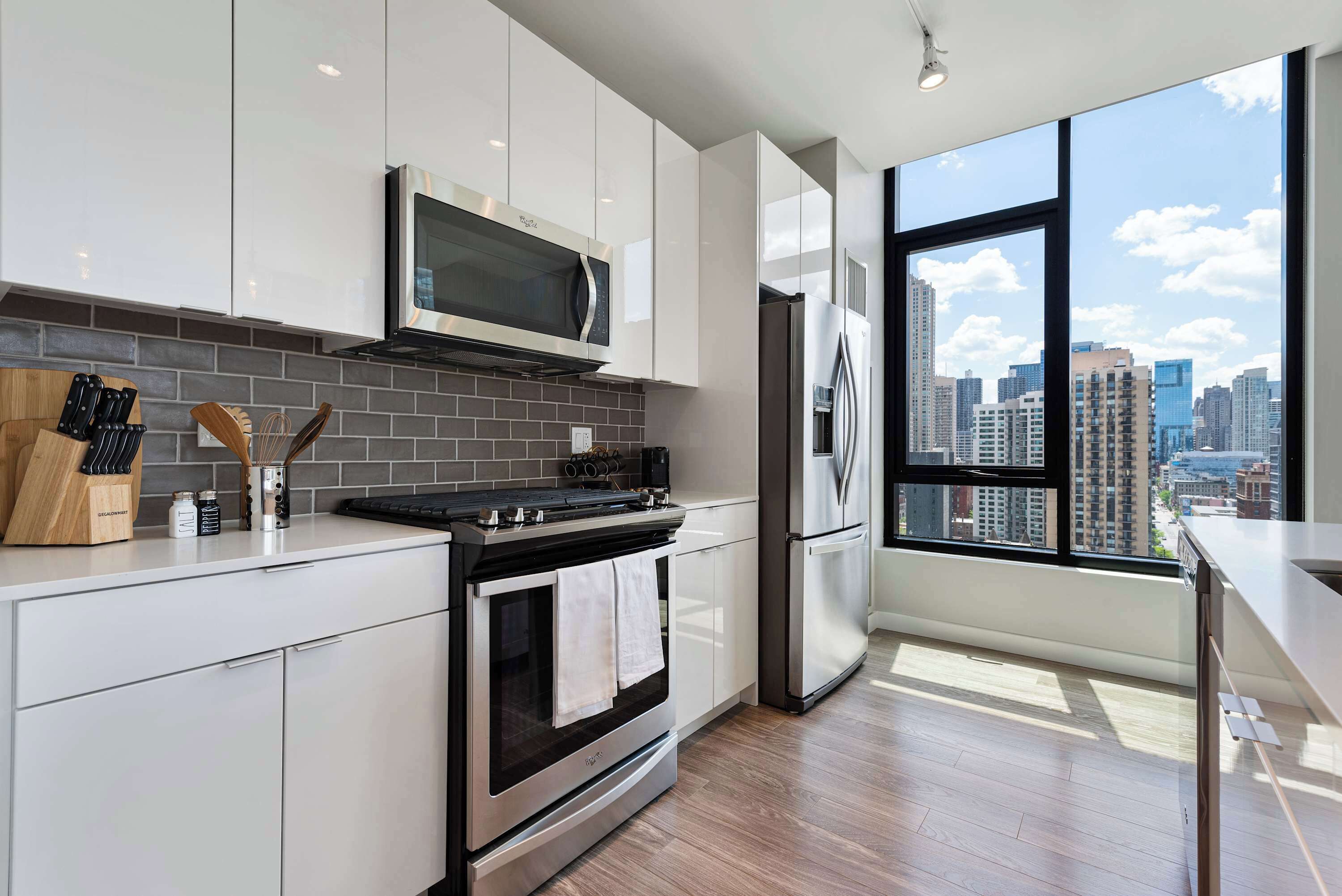 A modern kitchen with white cabinets, a stainless steel range, a microwave, and a refrigerator. There's a large window offering a city view.