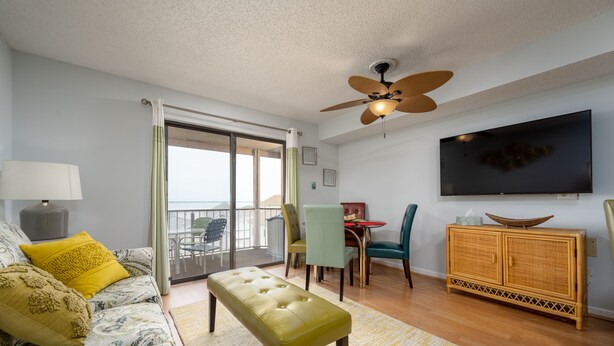 A living room with a sofa, bench, dining table, chairs, a TV, and a balcony with a view. - Contemporary, clean 1-bedroom mid-town condo with bay view and 1 block to beach. (Ocean City)