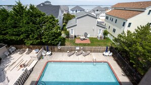 A rectangular swimming pool with a brick border, surrounded by a wooden deck with lounge chairs and a small patio area. - Contemporary, clean 1-bedroom mid-town condo with bay view and 1 block to beach. (Ocean City)
