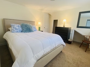 Een netjes opgemaakt bed met een witte dekbedovertrek en blauw gestreepte kussens, een klein nachtkastje met een lamp, een stoel en een open haard.
