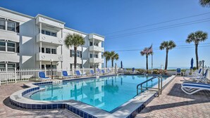 A swimming pool with a white building in the background, palm trees, and a clear blue sky. - Oceanside Ormond Beach Condo, Steps to Shore! (Ormond Beach)