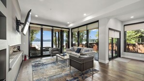 A modern living room with a large TV mounted on the wall, a sofa, a coffee table, and a view of the outdoors through large sliding glass doors. - Monthly 3BR Condo Rental on Whitefish Lake (Whitefish)