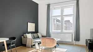 Ein modernes Wohnzimmer mit Sofa, Esstisch und Stuhl. Ein Fenster mit Vorhängen, ein Heizkörper und ein Bild an der Wand sind zu sehen.
