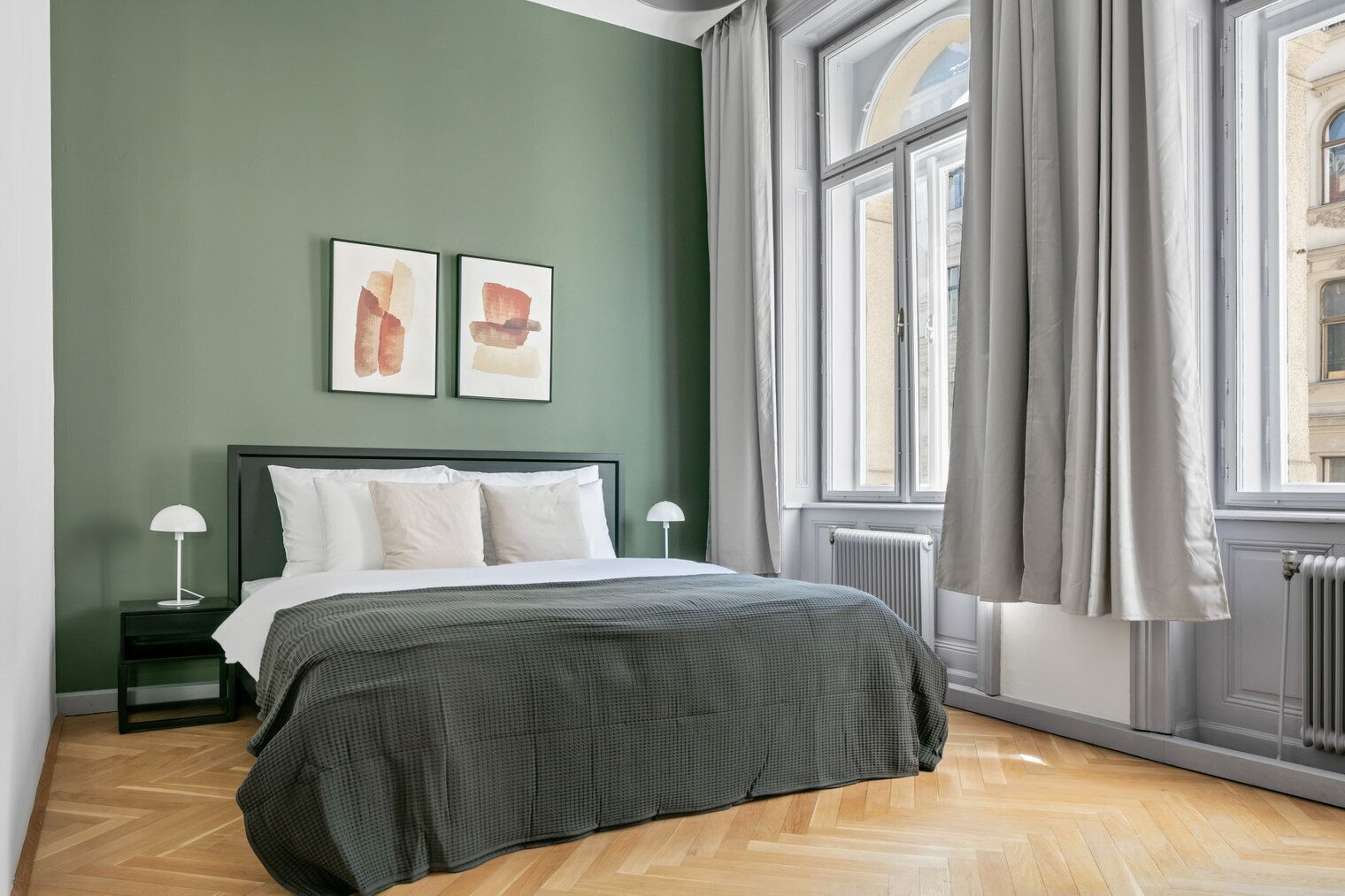 A bedroom with a bed, two bedside tables with lamps, a radiator, and a window with curtains.