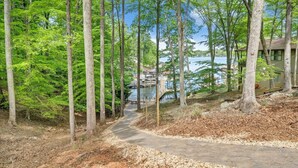 A forested area with a paved path leading to a dock by a lake, surrounded by tall trees and a building on the right. - Hokie Hideaway- Pet-friendly home with a fire-pit to enjoy with the family. (Huddleston)