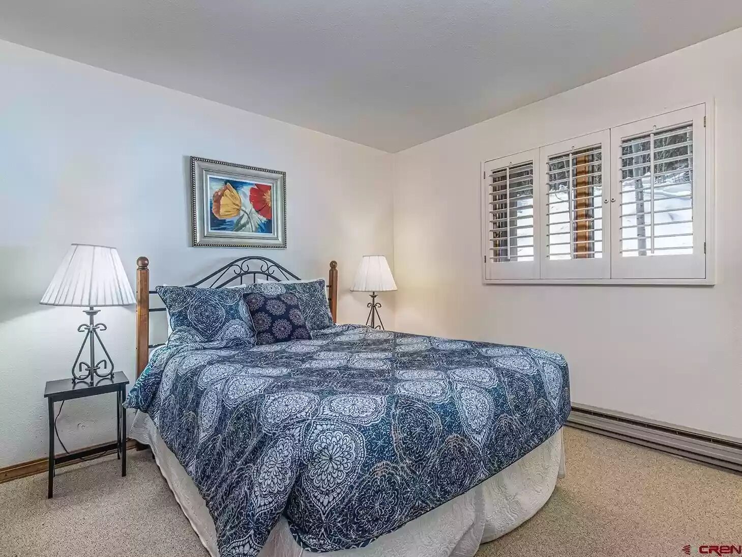 A bedroom with a bed, two bedside tables, a lamp, a picture on the wall, and a window with shutters.