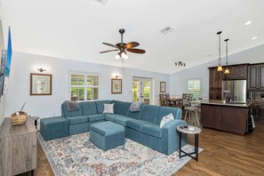 A spacious living area with a sectional sofa, a patterned rug, a ceiling fan, and a kitchen area with wooden cabinets and a stainless steel refrigerator.