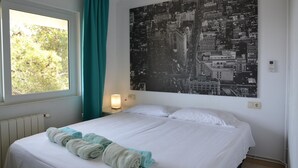 Et hotelværelse med en stor seng, et vindue med gardiner, en vægmonteret lampe og et bybillede som plakat.