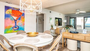 Un coin repas avec une table ronde, des chaises en osier, un ventilateur de plafond blanc et un tableau représentant un verre à cocktail.