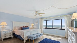 Ein Schlafzimmer mit Bett, Nachttischen, einer Bank, einem Deckenventilator und einem großen Fenster mit Blick auf den Strand.