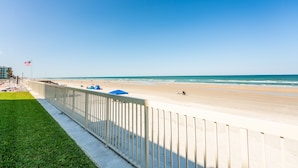A beach with a white fence, a grassy area, and a flag. - Castle Reef 132  Just Off the Sand (New Smyrna Beach)