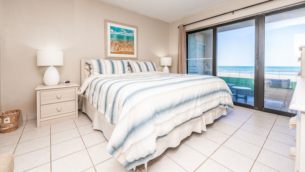 A bedroom with a bed, two bedside tables, a nightstand, a large window with a view of the sea, and a balcony. - Castle Reef 132  Just Off the Sand (New Smyrna Beach)