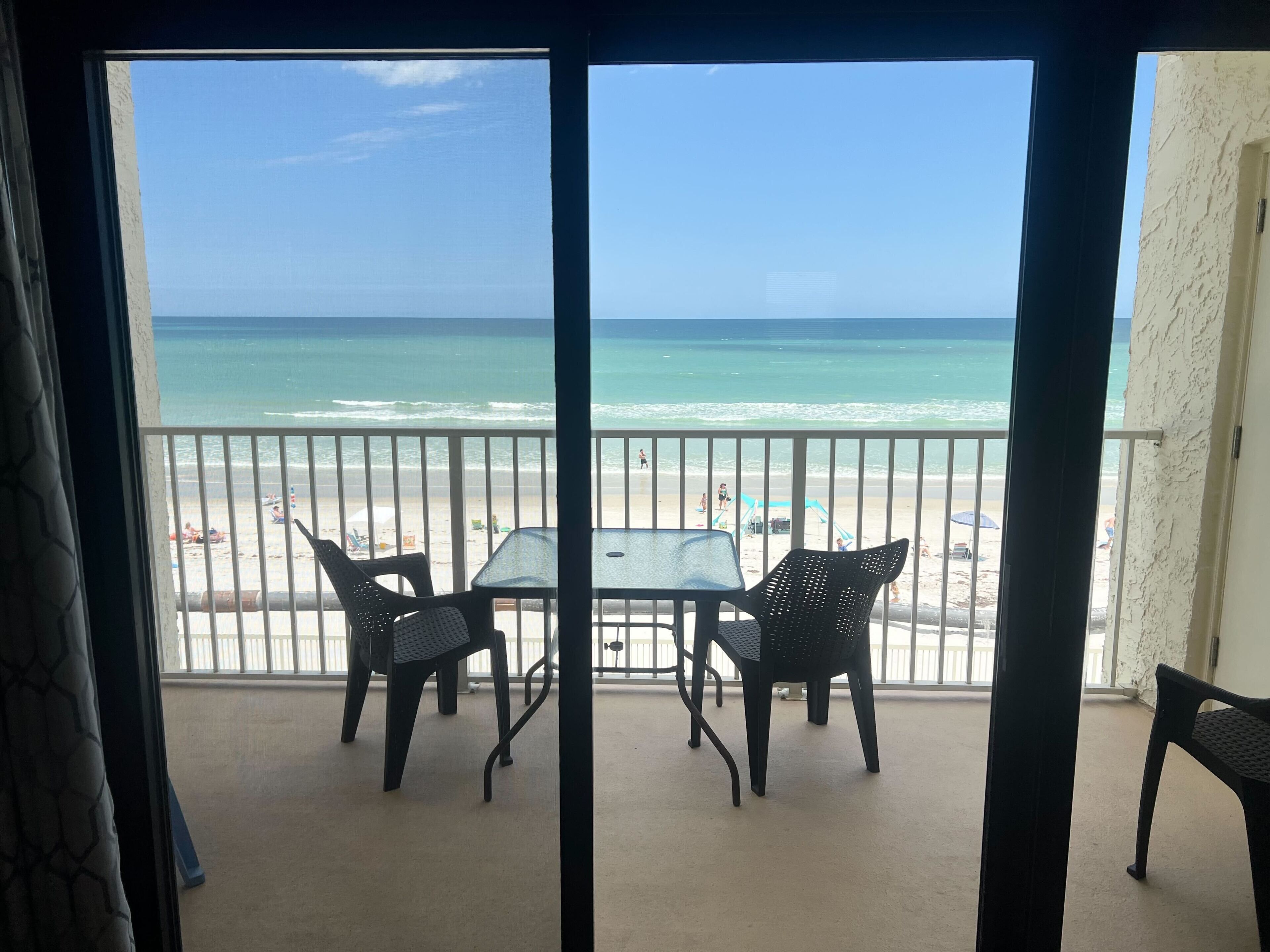 A balcony with a view of the beach, a table, and two chairs.