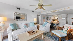 A living room with a white sofa, a wooden coffee table, and a blue patterned rug. There is a ceiling fan, a dining area with chairs, and a kitchen in the background.