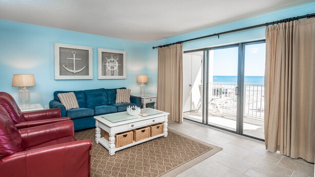 Una sala de estar con un sofá azul, sillas de cuero rojo, una mesa de centro blanca y vistas a la playa a través de amplios ventanales correderas.