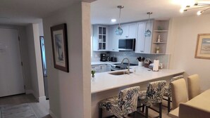 A modern kitchen with white cabinets, a stainless steel sink, and a built-in oven. There are two bar stools with patterned upholstery and a dining table with chairs. - Castle Reef 331 ultimate ocean views! (New Smyrna Beach)