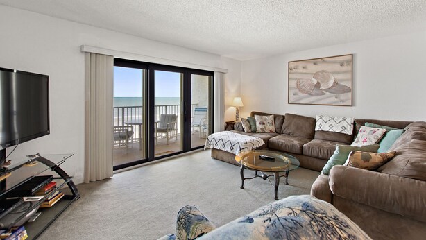 A spacious living room with a large flat-screen TV, comfortable seating, a coffee table, and a view of the ocean through a sliding glass door. - Castle Reef 331 ultimate ocean views! (New Smyrna Beach)