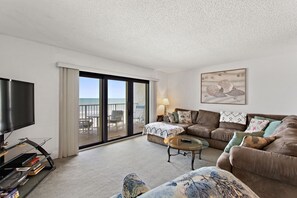 A spacious living room with a large flat-screen TV, comfortable seating, a coffee table, and a view of the ocean through a sliding glass door. - Castle Reef 331 ultimate ocean views! (New Smyrna Beach)