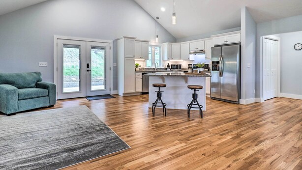 A modern kitchen with wooden flooring, white cabinets, a stainless steel refrigerator, and a bar area with stools. - Waynesville Smart Cabin w/ Hot Tub & Views! (Waynesville)
