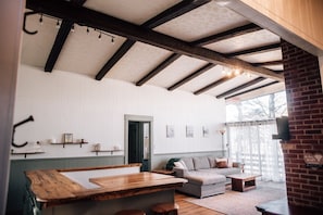 A spacious living area with a wooden bench, a sofa, and a coffee table. There is a door leading to another room, and a brick wall on one side.