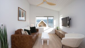 A modern living room with a brown sofa, white coffee table, and a white armchair. A ceiling fan is visible, and there is a large window with a view of a mountain and a wooden structure.