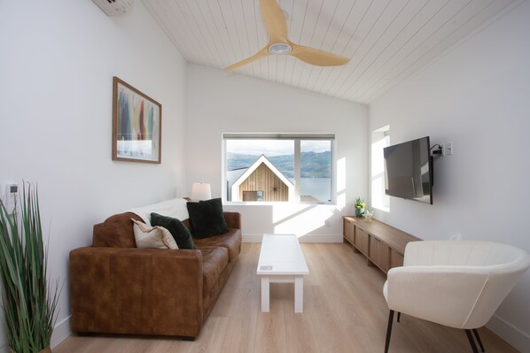 A modern living room with a brown sofa, white coffee table, and a white armchair. A ceiling fan is visible, and there is a large window with a view of a mountain and a wooden structure.