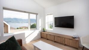 A modern living room with large windows, a flat-screen TV, a wooden bench, and a white coffee table.