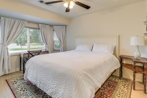 Ein Schlafzimmer mit einem großen Bett, einem Schreibtisch, einem Sessel, einem Deckenventilator, einem Fenster mit Vorhängen und einem Nachttisch mit einer Lampe.