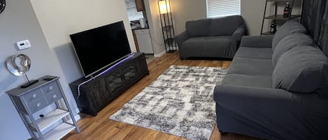 A living room with a flat-screen TV, grey sofa, wooden flooring, and a patterned rug.