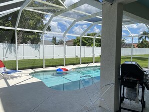 Une piscine extérieure couverte, entourée d’une clôture blanche, avec un barbecue et une chaise longue.