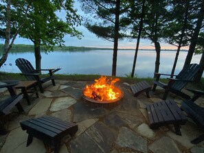 A stone fire pit surrounded by Adirondack chairs with a lake view.