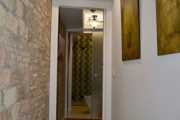 A hallway with a stone wall, a door leading to a room with a patterned wallpaper, and a light fixture above.