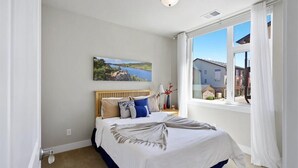 Un dormitorio con una cama grande, un cuadro de un paisaje, una ventana con cortinas y vistas a edificios.