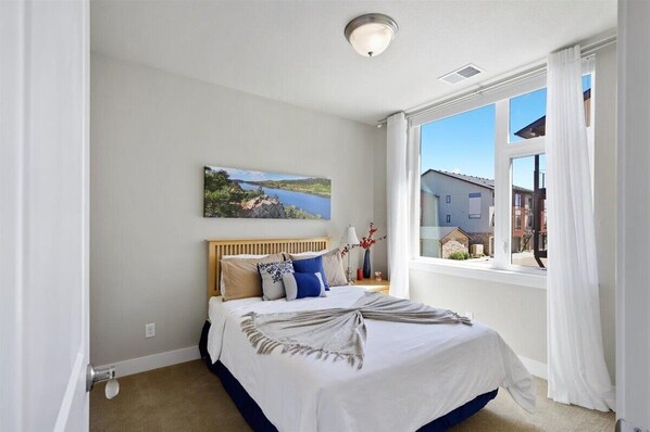 A bedroom with a large bed, a painting of a landscape, a window with curtains, and a view of buildings outside.