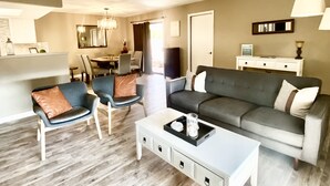 Een moderne woonkamer met een grijze bank, twee fauteuils, een salontafel en een eethoek op de achtergrond.