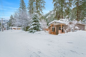 Una cabaña cubierta de nieve con fachada de madera y un porche, rodeada de árboles cargados de nieve y montañas al fondo.