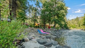 A riverside area with a rocky shore, a blue and pink lounge chair, and a wooden cabin in the background.
