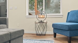 A living room with a blue armchair, a small side table with a lamp, a wooden floor, and a view of a deck through a window.