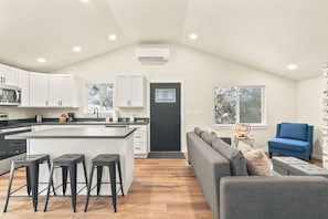 Una cocina moderna con armarios blancos, una isla central con taburetes y un sofá gris en una sala de estar con vistas a árboles.